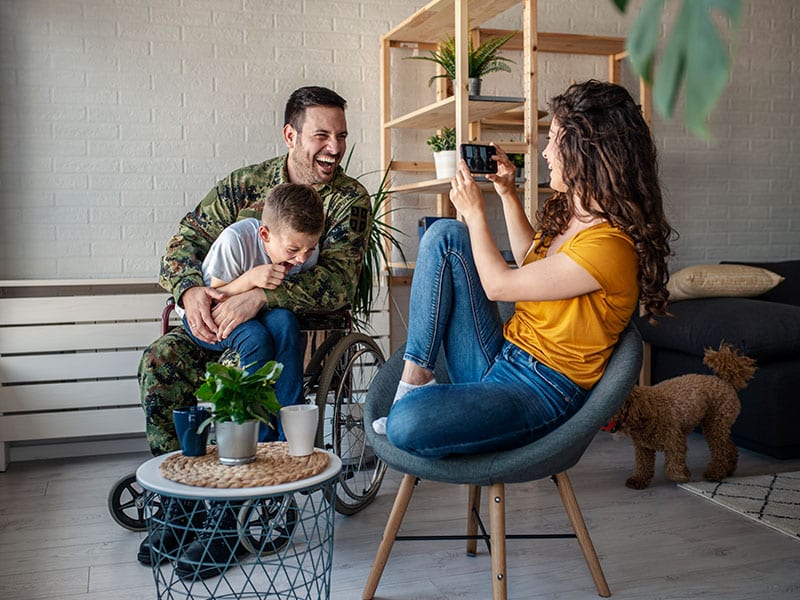 Smiling military parent in wheelchair hugging child while partner captures the moment at home